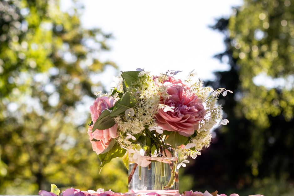 A delicate floral arrangement in a clear glass vase, featuring soft pink peonies with ruffled petals, accented by small white blossoms and lush green leaves. The bouquet is set outdoors on a sunny day