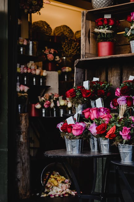 An indoor floral display featuring fresh pink and red roses arranged in small metallic buckets placed on a round black table. The arrangement showcases a variety of rose sizes with delicate, velvety p