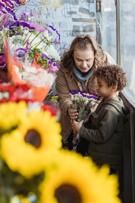 A young woman with light curly hair wearing a light brown jacket and a blue turtleneck is handing a small bouquet of purple and white flowers wrapped in clear plastic to a young boy with dark curly ha
