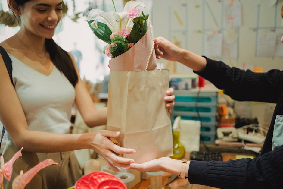 A young woman with long dark hair, wearing a sleeveless white top, is smiling as she receives a floral gift bag from a person dressed in black. The bouquet inside the paper bag features fresh white li
