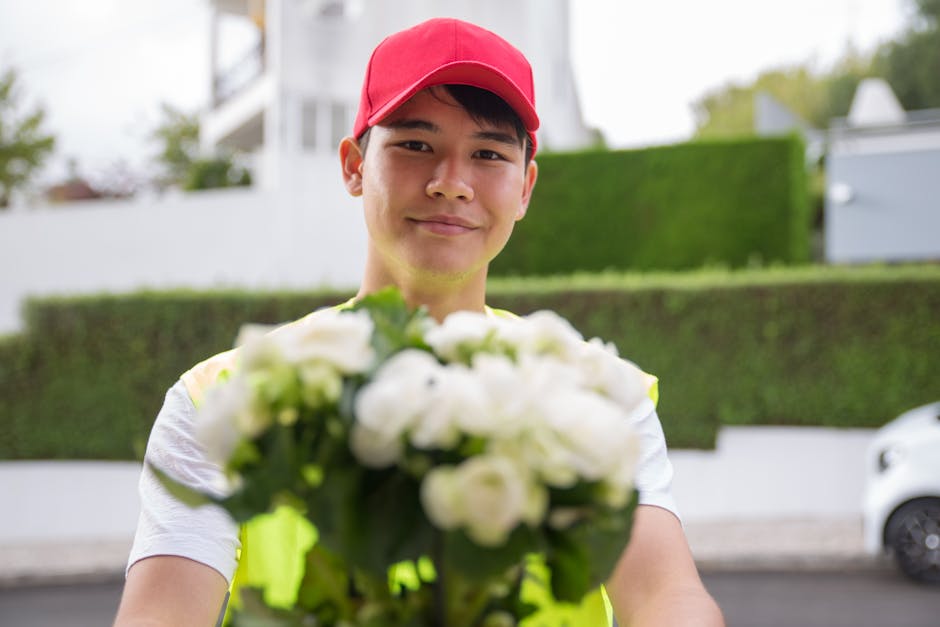 A delivery person, wearing a red cap and a high-visibility yellow vest, stands outdoors holding a fresh bouquet of white roses and green foliage, offering the floral gift towards the camera. The bouqu